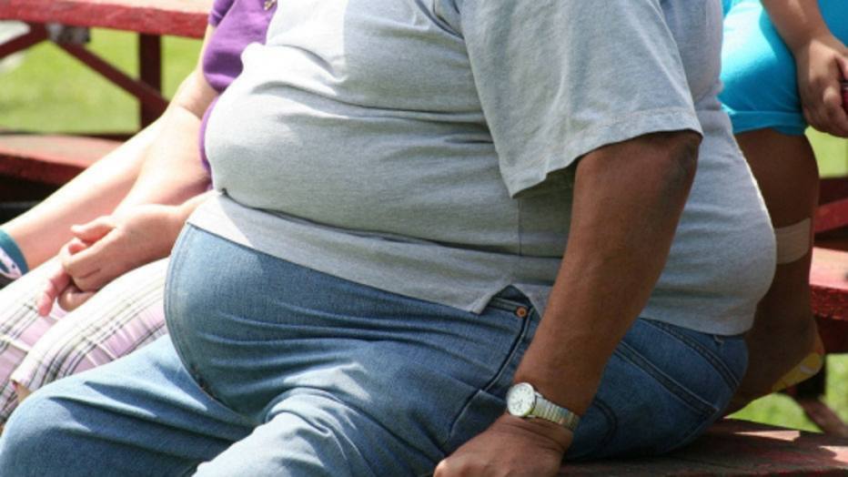 Uma pessoa obesa de calça jeans e blusa cinza sentada em um banco ao ar livre.