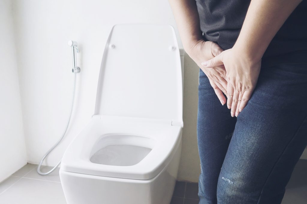 Incôntinência urinária. Mulher ao lado do vaso sanitário com a mão na bexiga, segurando a urina