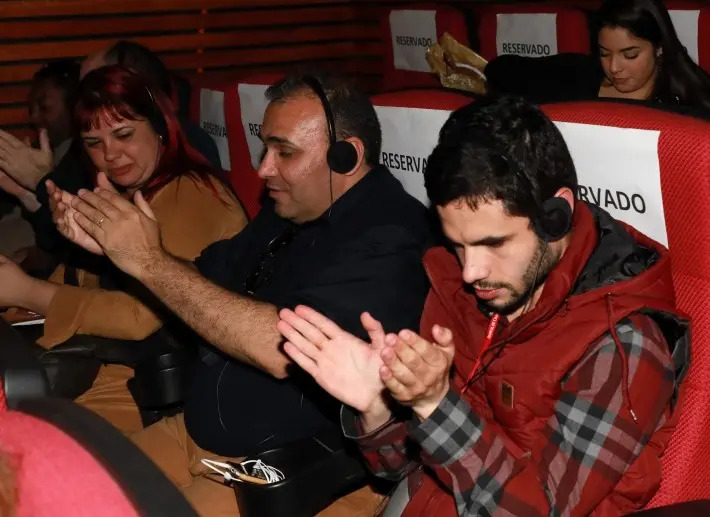 Dois homens cegos estão sentados em poltronas reservadas de uma sala de cinema. Eles usam fones de ouvido e aplaudem. (Foto: Reprodução)