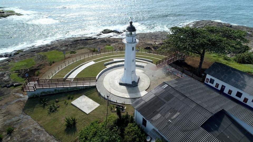 A imagem destaca um grande farol branco em frente a praia. É o Farol de Santa Luzia, em Vila Velha