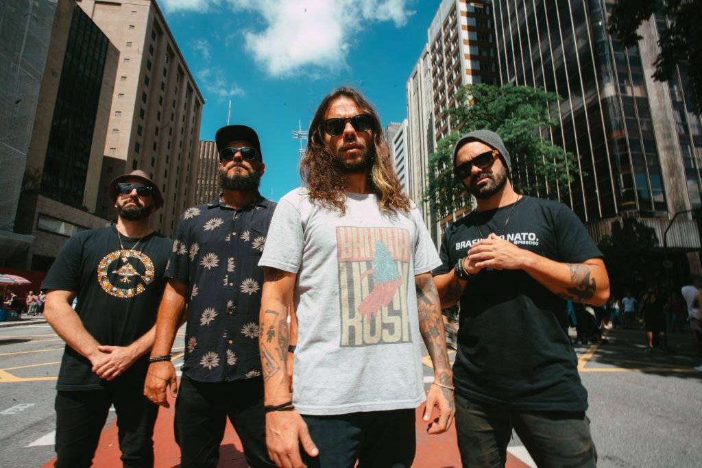 Banda Macucos posando pra foto em meio a prédios. Os quatro integrantes da banda.