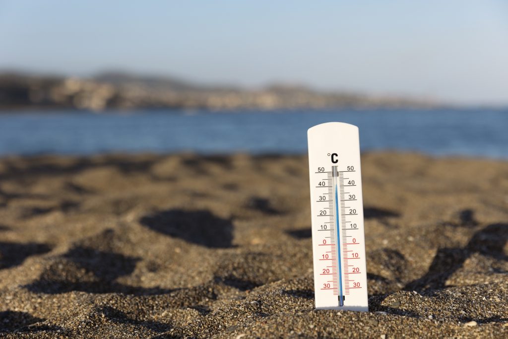 Termômetro afundado na areia com o mar ao fundo. A foto é para falar que está calor, mas tem previsão de chuva para o fim de semana