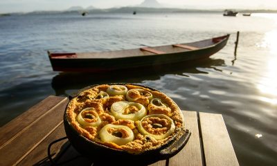 Torta capixaba na panela de barro em cima de uma mesa e o mar ao fundo com um barco na Ilha das Caieiras, em Vitória
