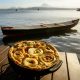 Torta capixaba na panela de barro em cima de uma mesa e o mar ao fundo com um barco na Ilha das Caieiras, em Vitória
