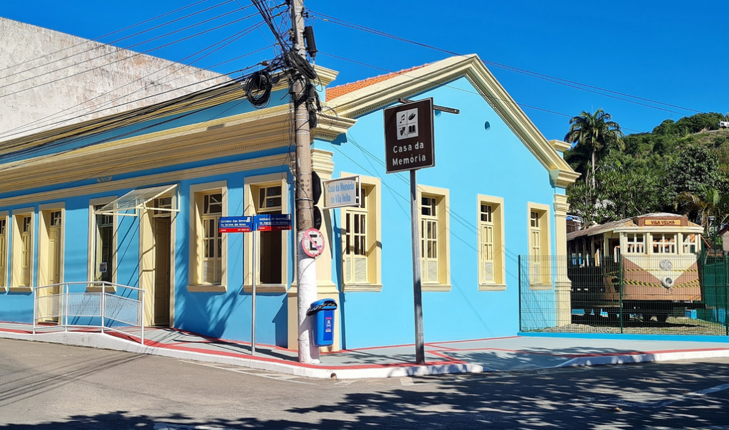 A imagem apresenta um casa azul com janelas na cor creme, uma placa para registrar o local e um bonde na parte de trás, um registro histórico de Vila Velha