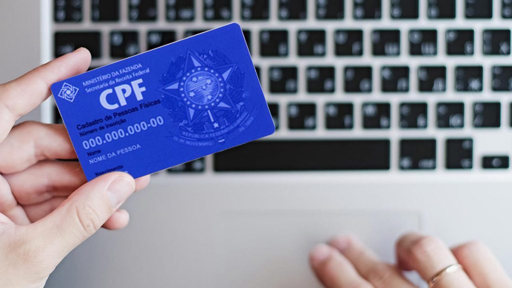 CPF na mão de uma pessoa e um notebook ao fundo.
