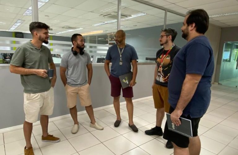 Cinco homens conversando em pé no corredor do trabalho. Todos eles utilizam bermudas na altura do joelho e camisetas. Alguns estão utilizando crachá e segurando caderno ou livro.