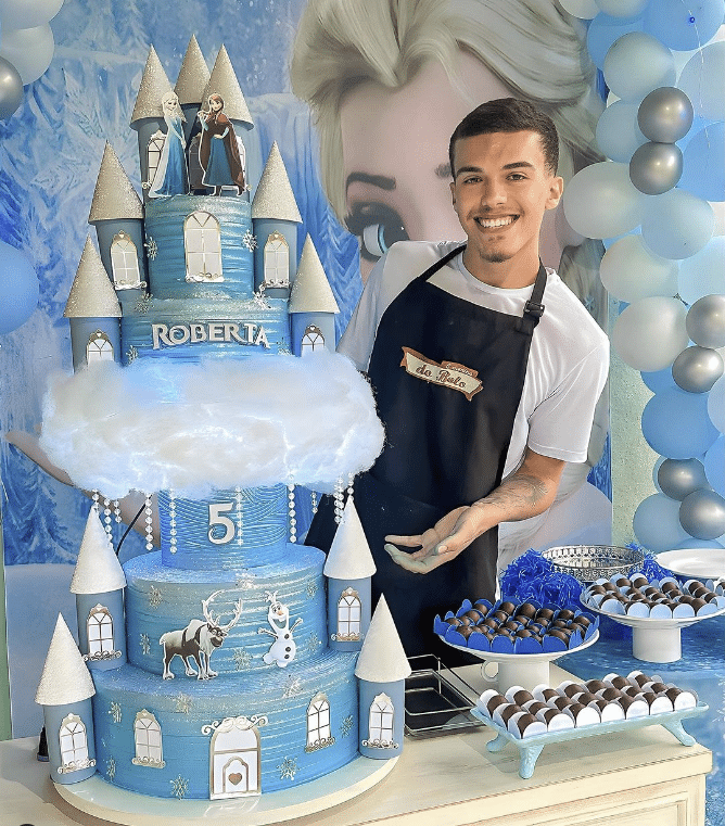 Bolo com tema Frozen todo azul e branco de cinco andares, com as torres do castelo. O confeito ao lado, com uma mesa de docinhos. Festa de criança