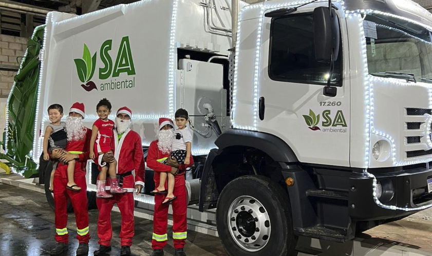 Caminhão de limpeza urbana da SA Ambiental decorado com luzes para o Natal. Servidores na frente vestidos de papais noeis com uma criança no colo