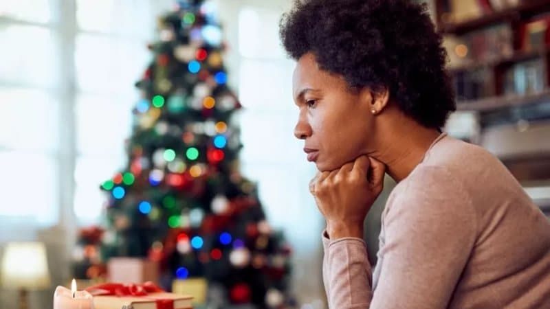 Na foto uma mulher negra com cara de pensativa aparece em primeiro plano. Ao fundo, de forma desfocada, uma árvore de natal