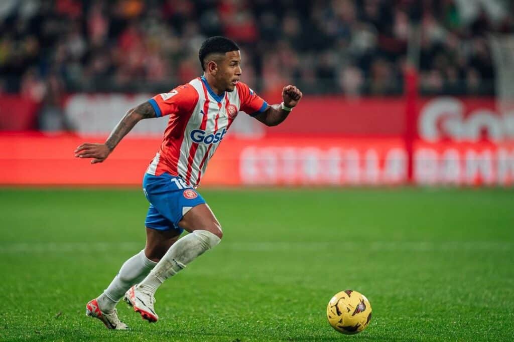 Na foto, um homem de pele preta vestindo uniforme de futebol nas cores branca, vermelha e azul, corre com a bola amarela pelo campo