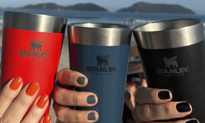 Três copos Stanley brindando na praia com mãos de mulheres.