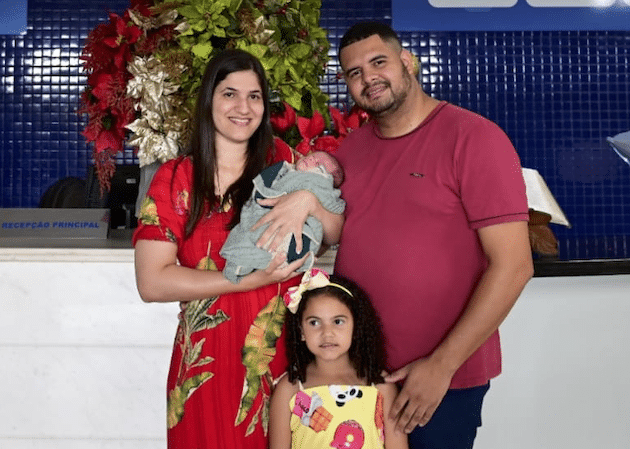 Pai e Mãe do sêxtuplos com o bebê no colo e a filha mais velha. Eles estão no hospital se preparando para levar o Henry para casa