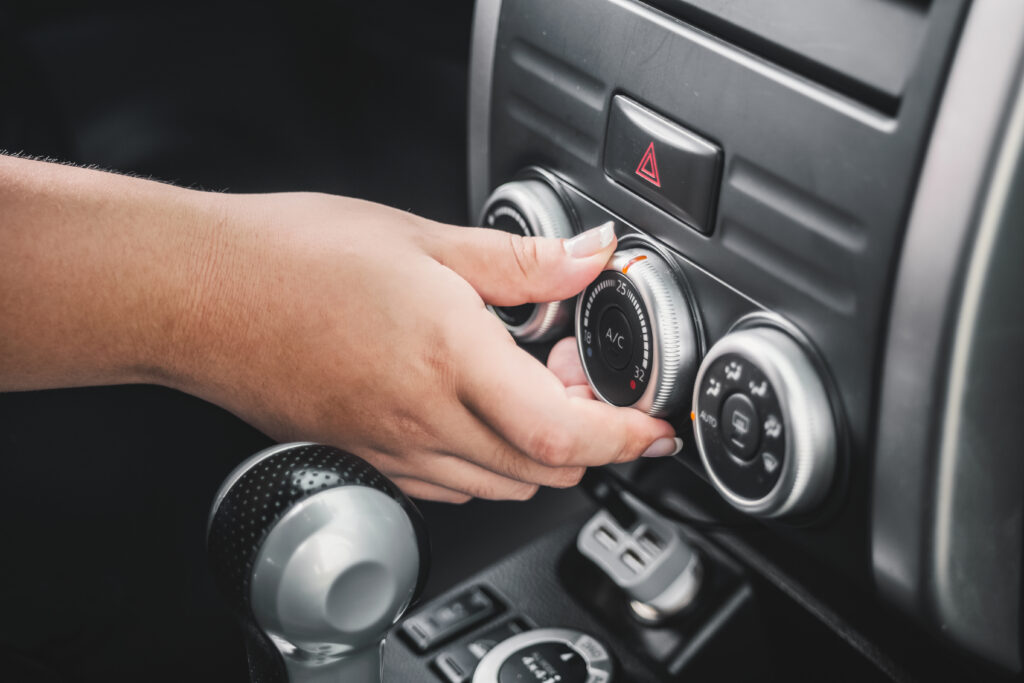 Na imagem aparece a mão de uma mulher mexendo no botão de controle do ar-condicionado de um carro