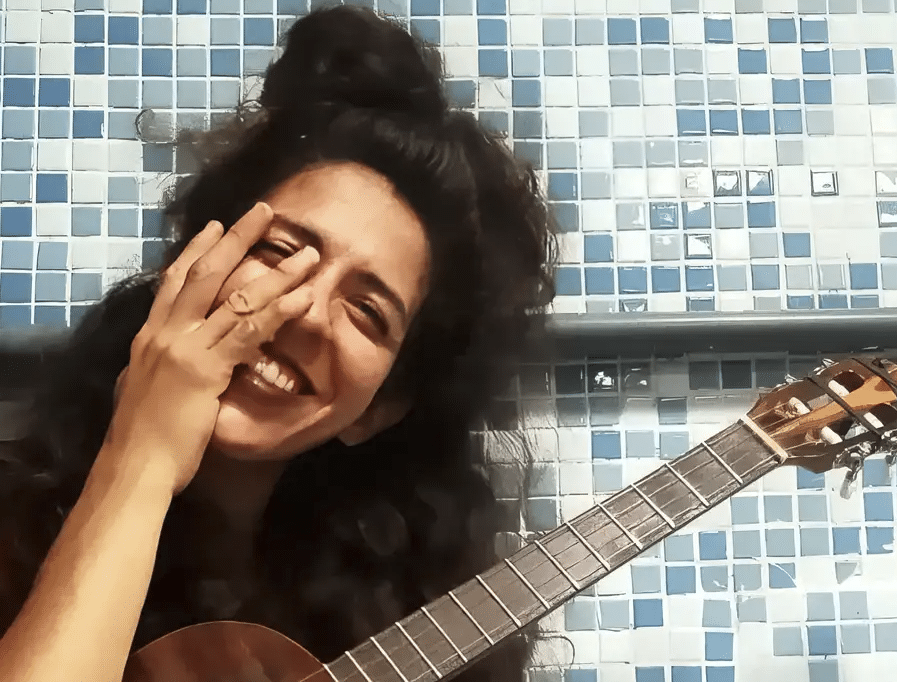 artista venezuelana com a mão no rosto e abraçada com um violão