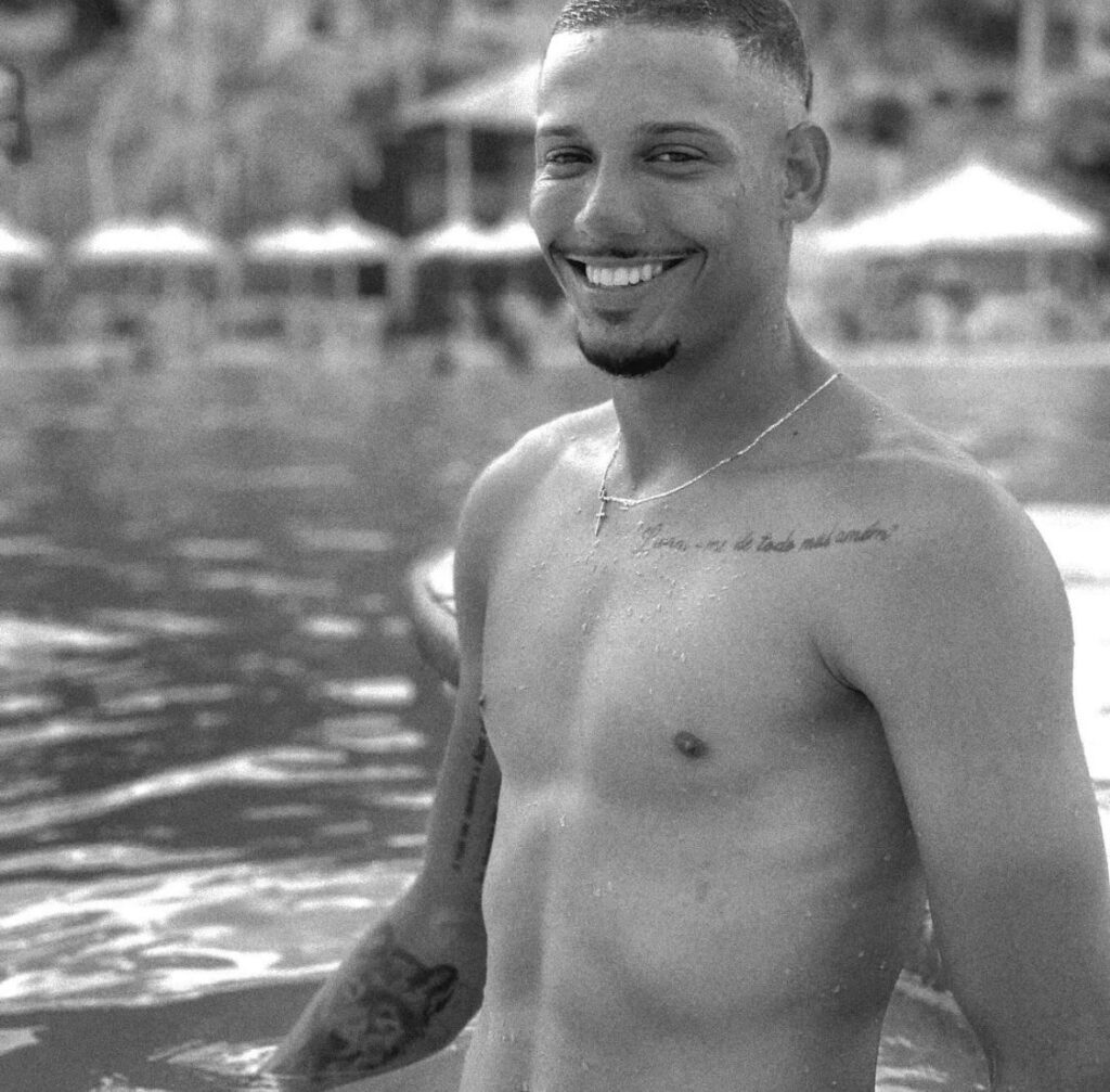 Caio de Oliveira em preto e branco, ao fundo é possível ver uma piscina e barracas para proteger do sol