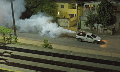 Carro do fumacê passando por um bairro da Serra. Da pra ver a fumaça saindo. O carro passando em uma rua cheia de casas