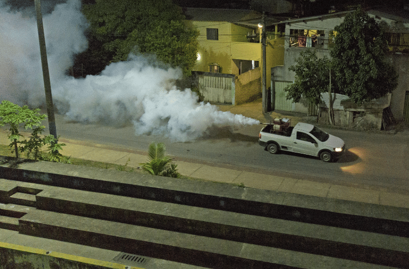 Carro do fumacê passando por um bairro da Serra. Da pra ver a fumaça saindo. O carro passando em uma rua cheia de casas