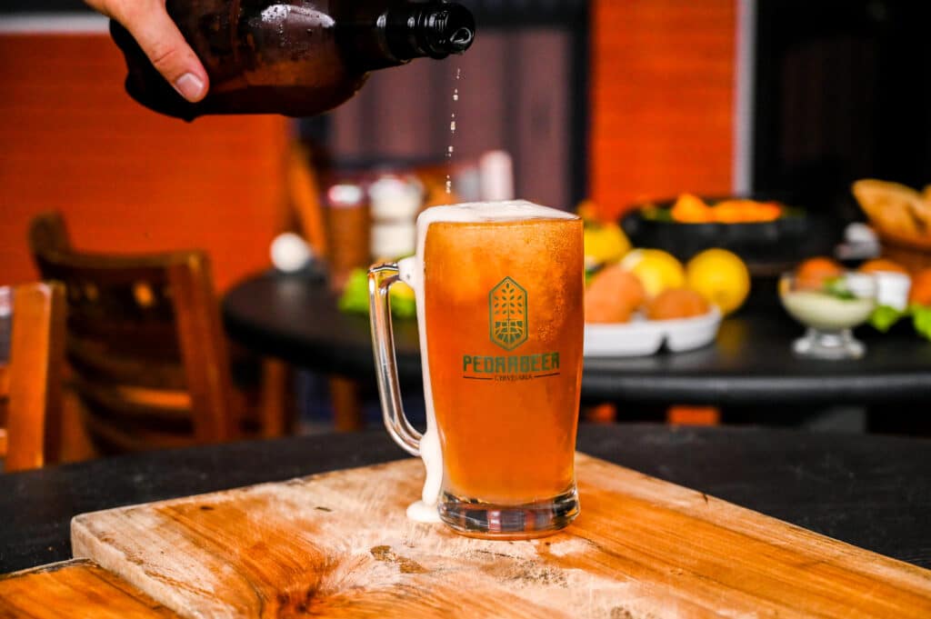 Cerveja artesanal Pedra Azul sendo despejada em uma caneca sobre uma mesa e ao fundo diversos petiscos