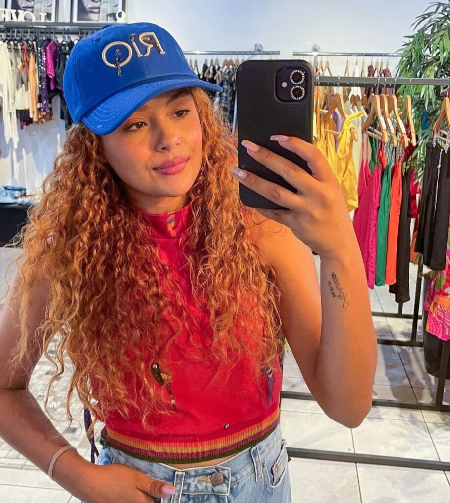 Lidia Amelia de Almeida Silva posando em uma loja de roupa com boné, blusa laranja e short jeans. Ao fundo araras cheias de roupas. A influenciadora morreu em um acidente de trânsito