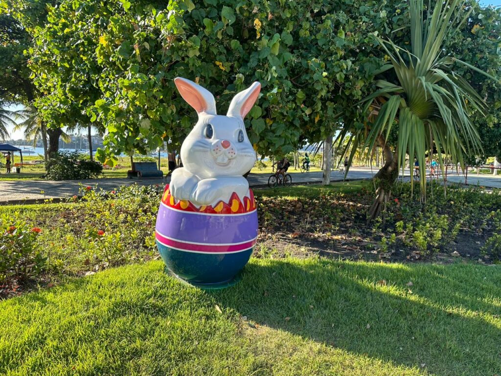 Um coelhinho saindo de um ovo colorido colocado em cima da grama. A ornamentação faz parte da Vila da Páscoa de Vitória, localizada na Praça dos Desejos