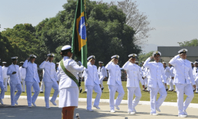 Oficiais da Marinha do Brasil enfileirados. Uma oficial com a bandeira do Brasil. Todos vestidos de branco. A Marinha do Brasil está com as inscrições abertas para concursos públicos