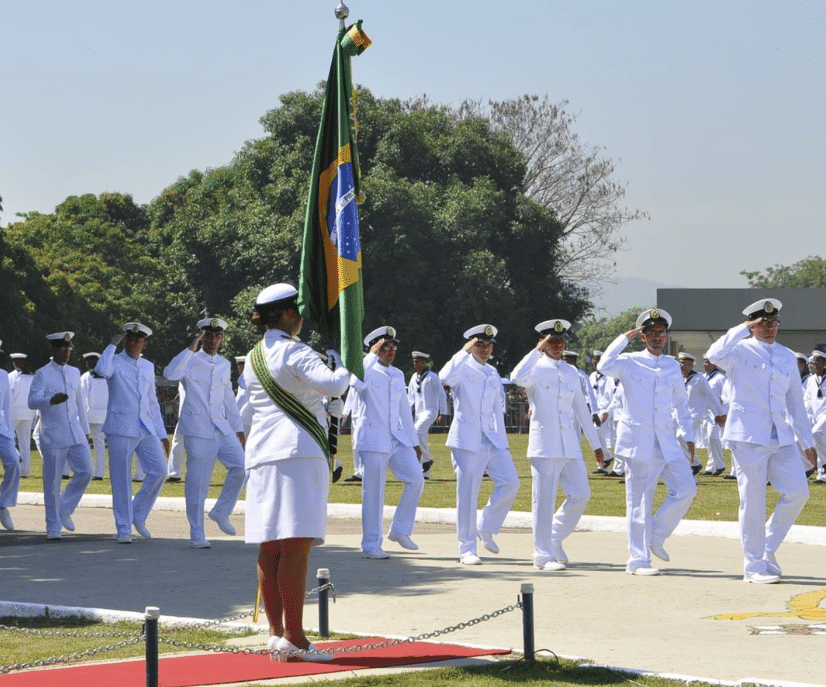 Oficiais da Marinha do Brasil enfileirados. Uma oficial com a bandeira do Brasil. Todos vestidos de branco. A Marinha do Brasil está com as inscrições abertas para concursos públicos
