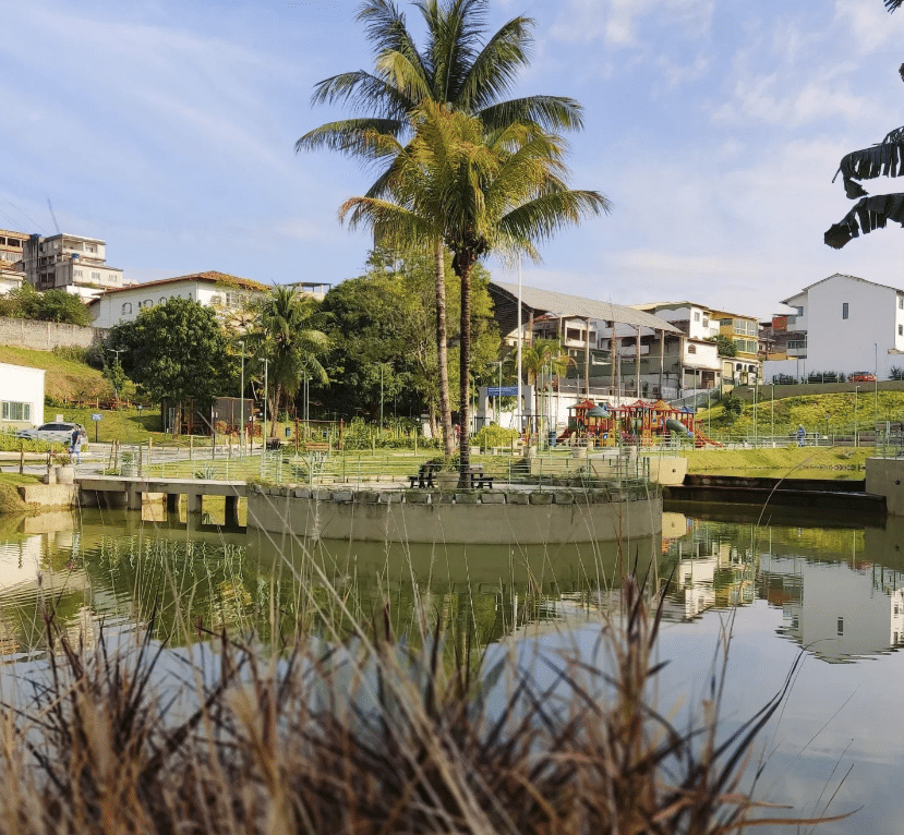 Um lago com muito verde no Parque da Biquinha, em Cariacica. Ao fundo tem um coqueiro e um parquinho para crianças