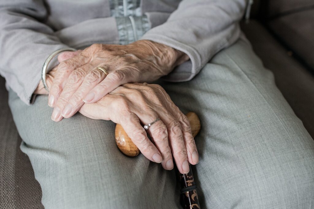 Mãos de um idoso repousando no colo segurando uma bengala que sugere que tem Parkinson