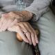 Mãos de um idoso repousando no colo segurando uma bengala que sugere que tem Parkinson
