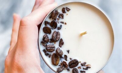 Uma mão segurando uma vela com cheiro de café. Para identificar o cheiro, tem sementes de café em cima