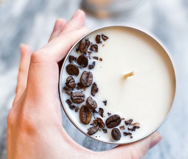 Uma mão segurando uma vela com cheiro de café. Para identificar o cheiro, tem sementes de café em cima. Os produtos serão lançados na ExpoSul Rural