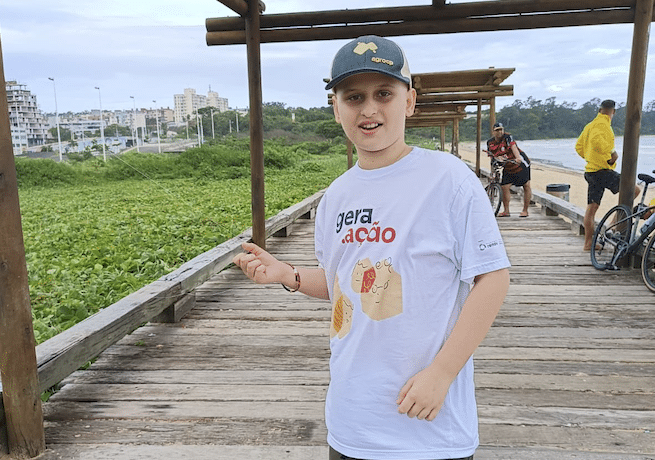 Victor Bonomo Trés, 12 anos, de boné, camisa e bermuda. Ele posa em uma estrutura de madeira próximo à praia. A família do menino lançou uma vaquinha online para custear o tratamento de câncer
