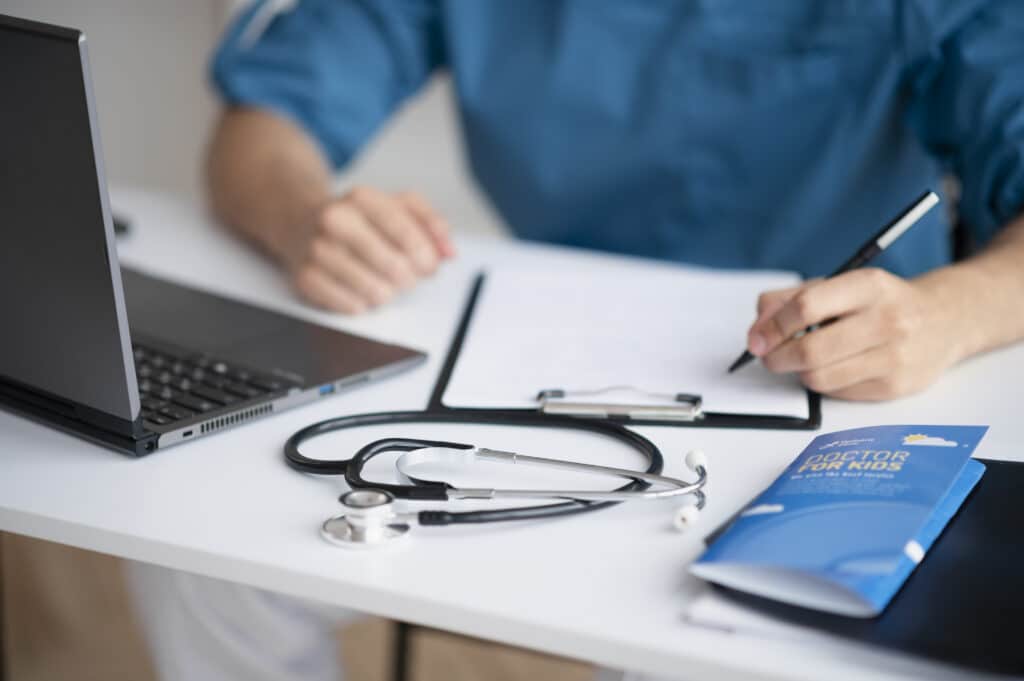 Médico em atendimento na mesa de trabalho com um estetoscópio, notebook e prontuário. Ele atende por planos de saúde