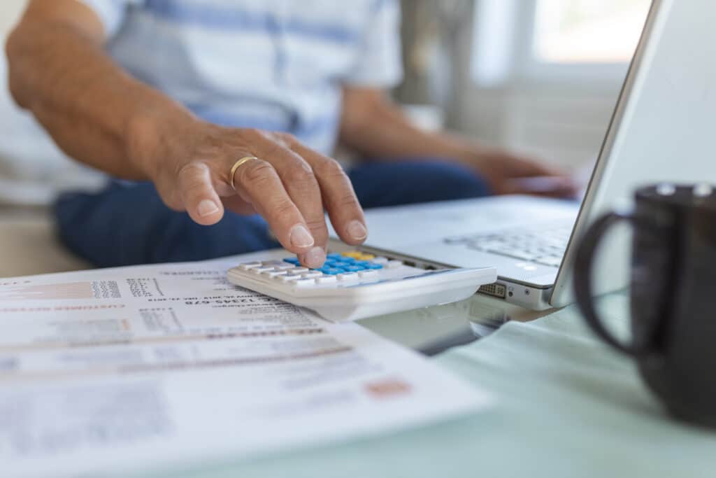 Um homem fazendo conta em uma calculadora com diversos papéis e um computador sobre dívidas