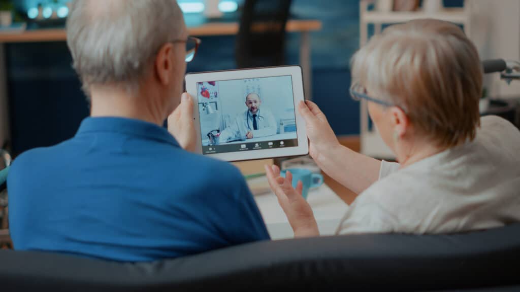 Um casal de idosos sentados com um tablete na mão passando por uma consulta de telemedicina