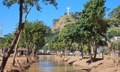 Um rio com árvores e ao fundo o Cristo Redentor de Mimoso do Sul