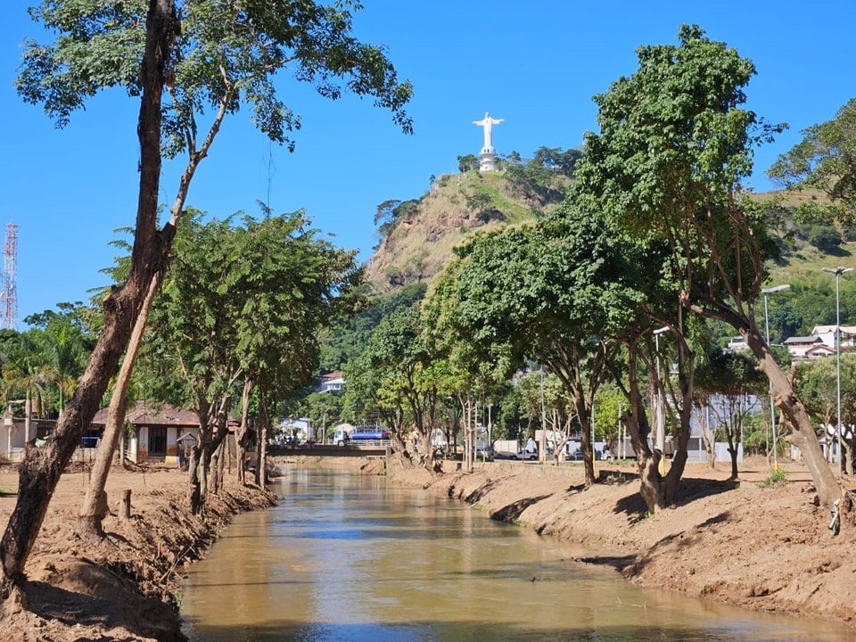 Um rio com árvores e ao fundo o Cristo Redentor de Mimoso do Sul