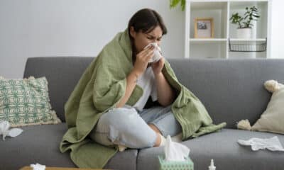 Uma mulher no sofá espirrando devido a um resfriado. Ela está enrolada em uma coberta verde