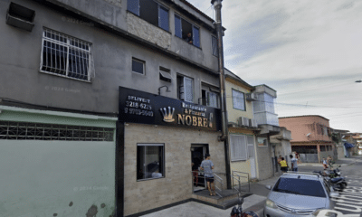 Imagem mostrando a fachada do Restaurante e Pizzaria Nobre, localizado em uma rua movimentada. O estabelecimento tem um letreiro visível com o nome e o logotipo da coroa. Algumas pessoas estão próximas à entrada, e há veículos estacionados ao longo da calçada. A rua apresenta um ambiente urbano típico, com prédios residenciais e comerciais ao redor.