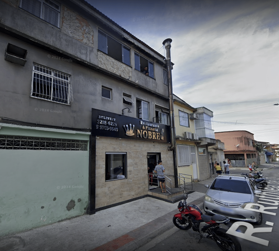 Imagem mostrando a fachada do Restaurante e Pizzaria Nobre, localizado em uma rua movimentada. O estabelecimento tem um letreiro visível com o nome e o logotipo da coroa. Algumas pessoas estão próximas à entrada, e há veículos estacionados ao longo da calçada. A rua apresenta um ambiente urbano típico, com prédios residenciais e comerciais ao redor.