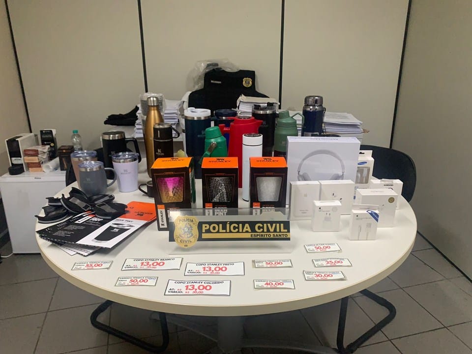 Mesa com vários produtos falsificados dispostos atrás de uma placa da Polícia Civil.