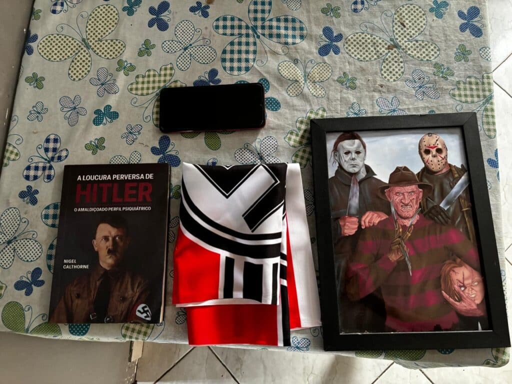 A imagem apresenta os materiais apreendidos pela Polícia Civil do Espírito Santo (PCES) durante uma investigação. Sobre uma mesa, estão dispostos um livro com o título "A Loucura Perversa de Hitler", com uma foto de Adolf Hitler na capa, uma bandeira com símbolo alusivo ao nazismo, um celular e um quadro com ilustrações de três personagens de filmes de terror, segurando facas: Michael Myers, Freddy Krueger e Jason Voorhees. Esses itens fazem parte das evidências recolhidas em um caso envolvendo um adolescente investigado por participação em grupos de ideologia nazista.