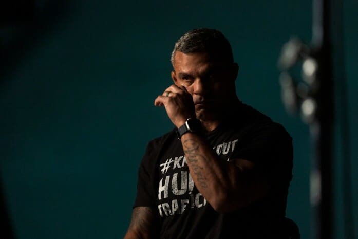 Na fotografia, o lutador Vitor Belfort aparece em um momento introspectivo, com expressão séria e postura contemplativa, enquanto enxuga uma lágrima. Ele veste uma camiseta preta com a frase "#Knockout Human Trafficking" (Nocaute no Tráfico Humano), evidenciando seu envolvimento em campanhas de conscientização sobre causas sociais. O ambiente está escuro, com iluminação suave, ressaltando a emoção e a gravidade do momento, possivelmente relacionado ao desaparecimento de sua irmã, Priscila Belfort. A imagem capta um lado mais vulnerável do atleta, demonstrando o impacto pessoal que o caso teve em sua vida.