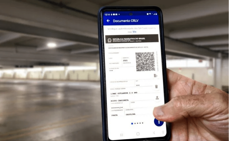 Mão de uma pessoa segurando um celular com o documento digital de licenciamento aberto. Ao fundo, um estacionamento coberto.