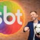 Thiago Leifert na frente da logo do SBT com uma bola de futebol na mão