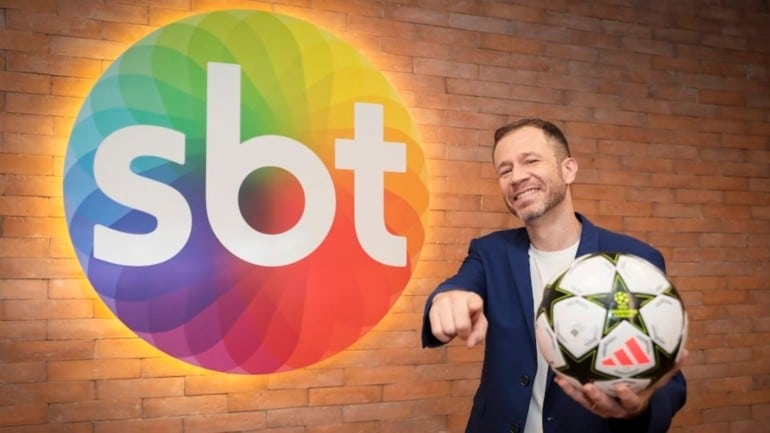 Thiago Leifert na frente da logo do SBT com uma bola de futebol na mão